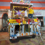 A towering, ornately decorated chariot (rath) adorned with vibrant fabrics and intricate carvings, surrounded by a sea of devotees pulling the ropes and celebrating in the streets during the annual Rath Yatra festival. The atmosphere is filled with energy, devotion, and the spirit of unity as participants from all walks of life join together in this grand tradition.